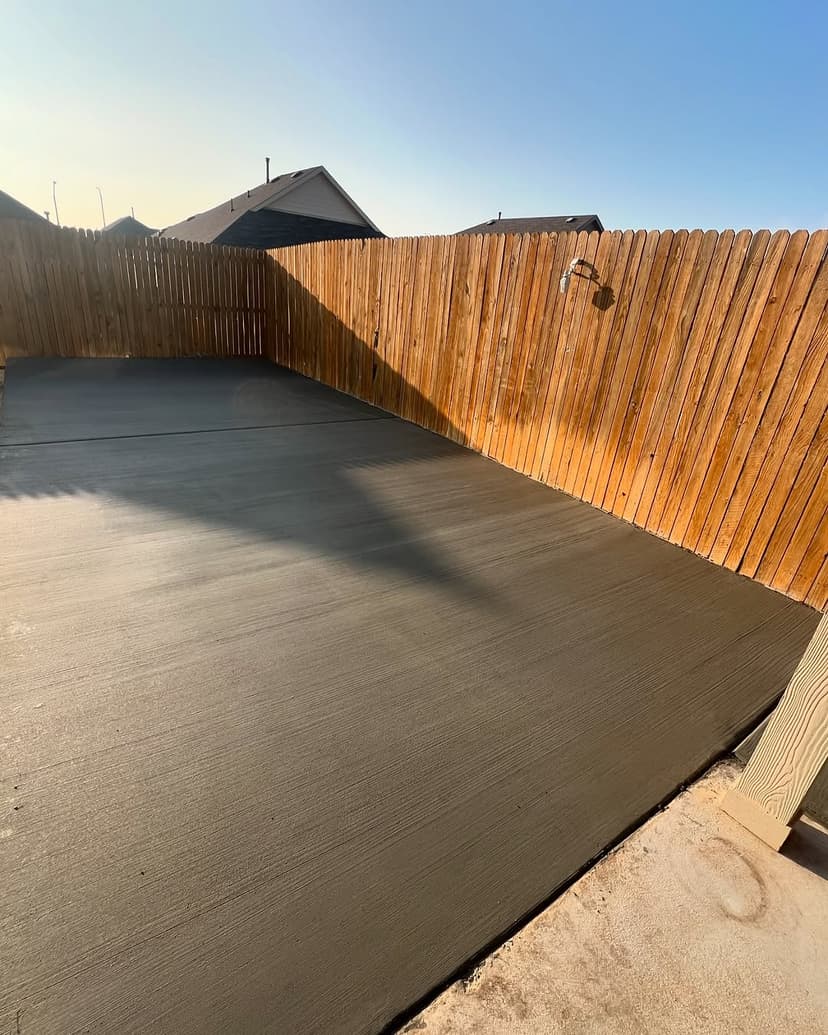 Freshly poured concrete surface in a fenced backyard with clear blue skies.