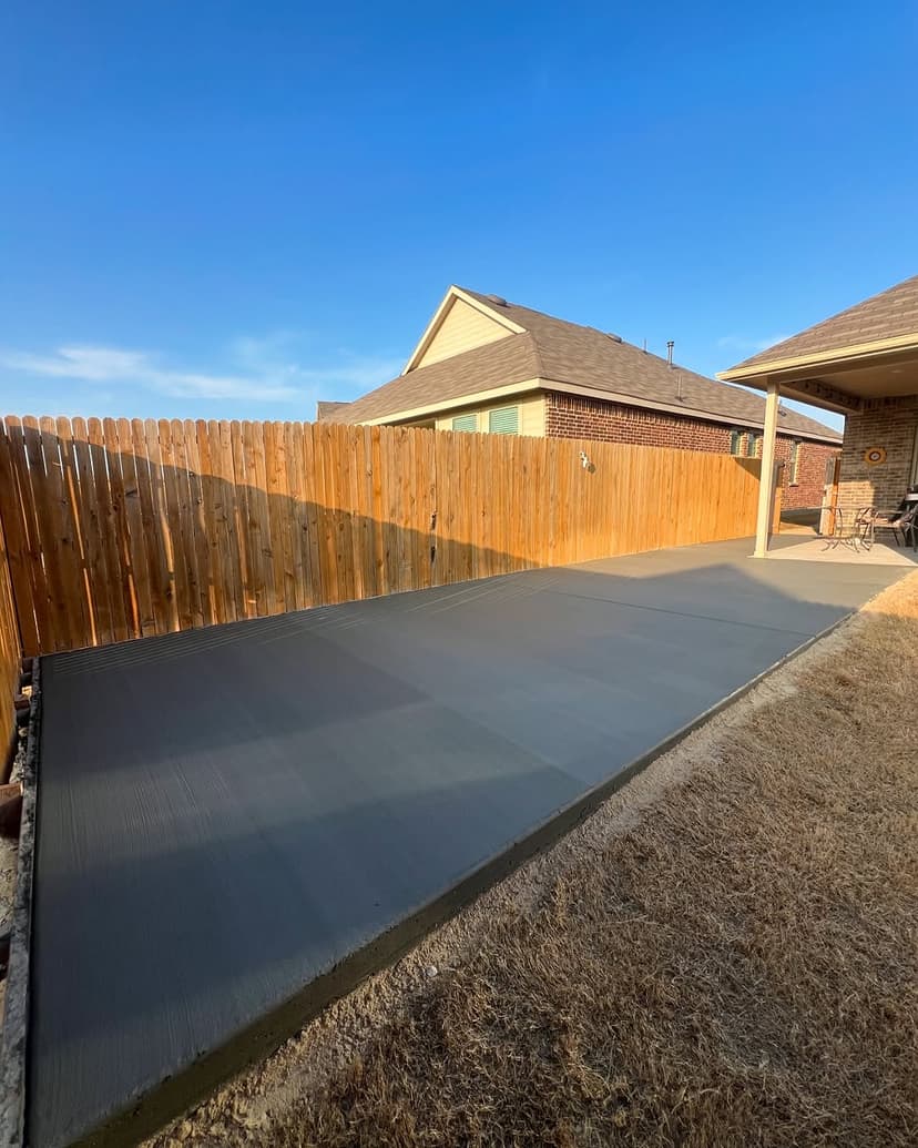 Freshly poured concrete slab in a residential backyard with wooden fencing and blue sky.