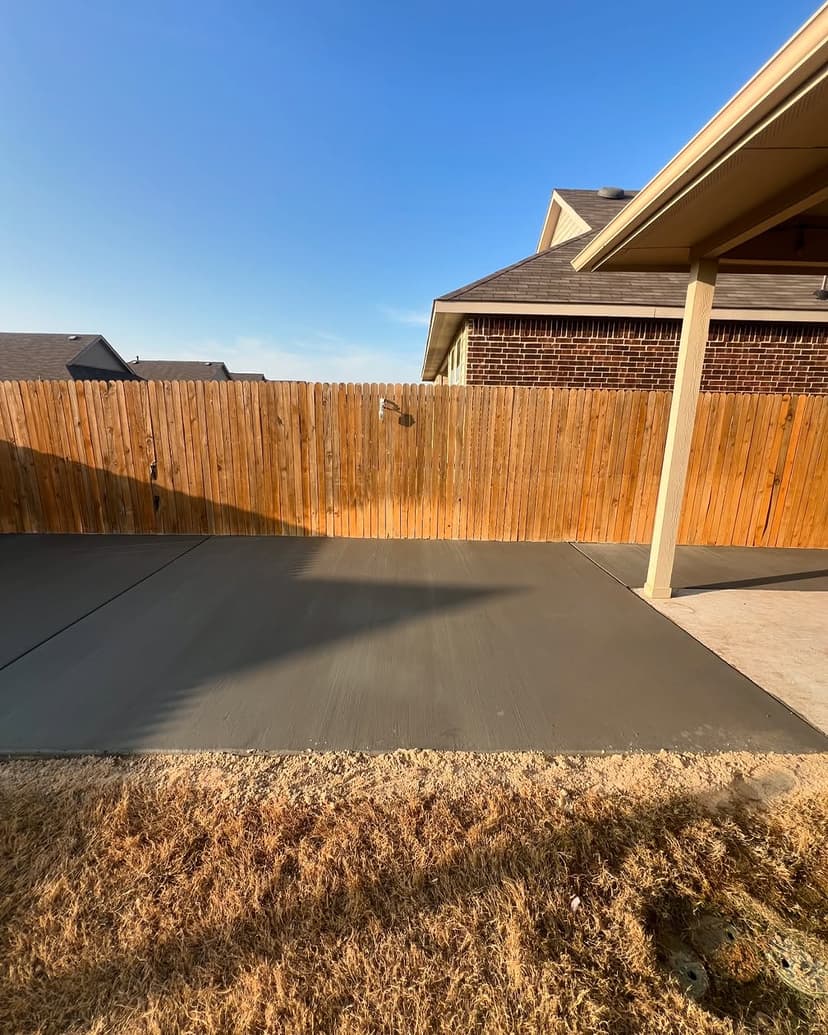 Freshly poured concrete patio next to a wooden fence under a clear blue sky.